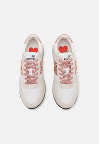 Sneakers featuring a white leather body, suede accents, and glossy gold studs. Pink glitter laces and a heart logo on the tongue.