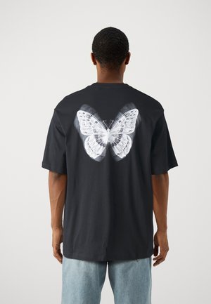 Mężczyzna w czarnej koszulce z dużym białym motylem na plecach, w jasnoniebieskich dżinsach, stojący na jednolitym tle.