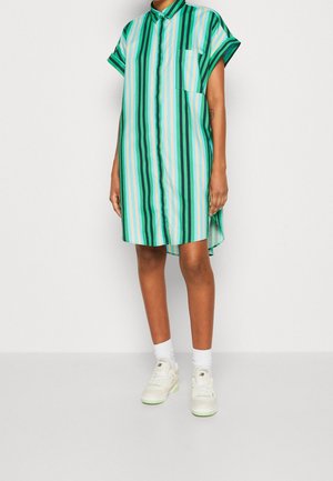 Osoba ubraną w krótki, zapinany na guziki sukienkę w paski w odcieniach zieleni i niebieskiego, do tego białe skarpetki i sportowe buty, stojąca na tle jednolitego tła.