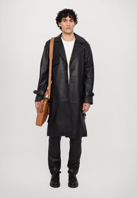 Svart läder trench coat med dubbelknäppt design, bred krage och justerbara ärmslut, kombinerad med svarta byxor och en brun axelväska.