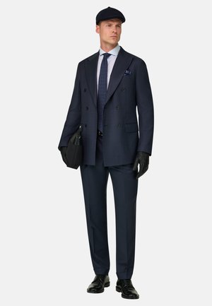 Homme portant un costume croisé bleu marine, cravate à motifs, chemise bleu clair, gants noirs, chaussures en cuir noir, casquette plate, tenant un sac noir.