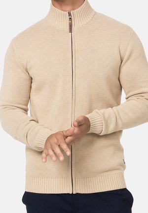 Homme portant un pull en maille beige zippé avec col et poignets côtelés, mains jointes devant, visible du cou à la taille.