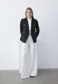 Kvinde stående op ad en hvid væg iført sort dobbeltknappet blazer, hvid flæset bluse, hvide bukser med vide ben og guldringøreringe.