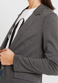 Šedý blazer z texturované pleteniny s klopovým límcem, přiléhavým střihem a dlouhými rukávy, nošený přes bílý tričko s potiskem.