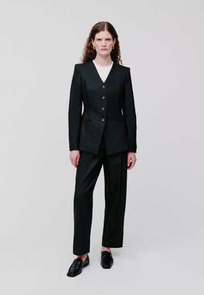 Schwarzer Anzug mit tailliertem Schnitt und strukturiertem Blazer mit tiefem V-Ausschnitt und drei Knöpfen, kombiniert mit geraden Hosen und schwarzen Loafers.