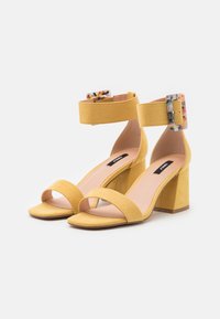 Keltaiset mokkasiinin korkokenkä sandaalit, joissa on leveä nilkkapanta värikkäällä soljella, avonainen varvasmuotoilu ja neliömäinen lohkokorko.