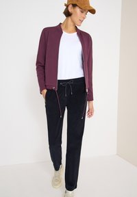 Burgundowa kurtka zapinana na zamek, biała koszulka, czarne spodnie z troczkami i beżowe sneakersy. Gładki materiał z kontrastowymi detalami i dopasowany krój.