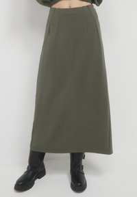Gonna verde scuro, lunga fino alla caviglia, con taglio dritto, caratterizzata da pieghe frontali e una superficie tessile liscia. Abbinata a stivali neri fino al ginocchio.