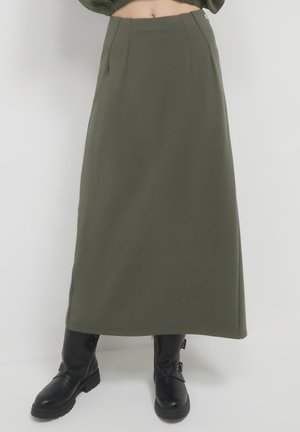 Gonna verde scuro, lunga fino alla caviglia, con taglio dritto, caratterizzata da pieghe frontali e una superficie tessile liscia. Abbinata a stivali neri fino al ginocchio.