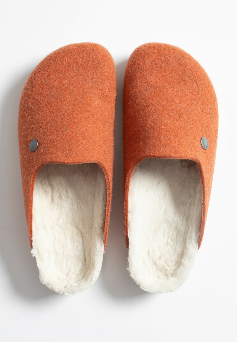 Zapatillas de fieltro naranjas con un suave forro de piel blanca, diseño de parte trasera abierta y un pequeño botón de acento gris en el lateral.