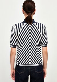 Pletené polo triko s námořnickým a bílým vzorem v chevronu, krátké rukávy a vroubkovaný límeček. V kombinaci s tmavými džínovými kalhotami.