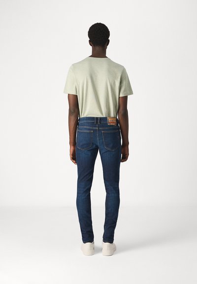 T-shirt vert clair associé à un jean slim bleu foncé. Le jean présente une patch visible à l'arrière avec des détails de label et une couture contrastante.