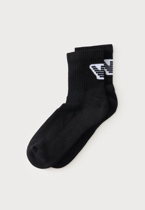 Paire de chaussettes noires type crew avec bords côtelés et détail de logo géométrique blanc sur les parties supérieures.