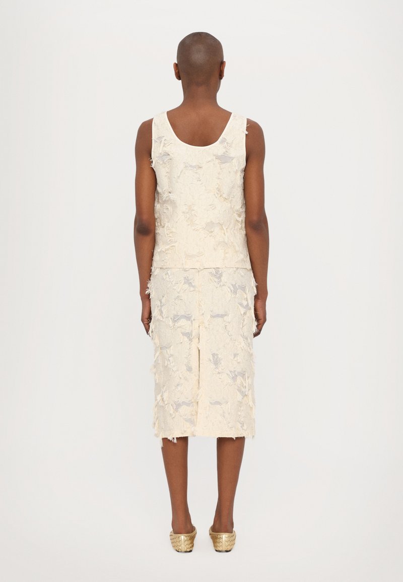 Ivoor mouwloos top met een textuur van bloemenpatroon, gecombineerd met een bijpassende knie-lange rok met ruwe randen en een subtiele glans. Gouden schoenen.