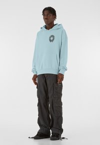 Lys blå hættetrøje med en frontlomme og cirkulært grafisk detalje. Kombineret med sorte cargo-bukser med flere lommer og snøre ved kanten.