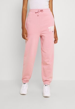 Person trägt pinke Jogginghose mit elastischen Bündchen und Kordelzug, weiße Sneakers und ein weißes Oberteil vor einfarbigem Hintergrund.