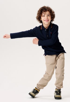 Polo à manches longues texturé bleu marine avec un col à trois boutons et des rayures multicolores ; pantalon beige fuselé ; baskets gris et noir.