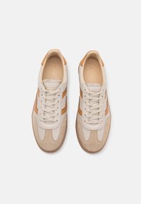 Béžové tenisky z kůže a semiše mají hnědé detaily, kulatou špičku, šněrování vpředu a gumovou podrážku s texturovaným vzorem.