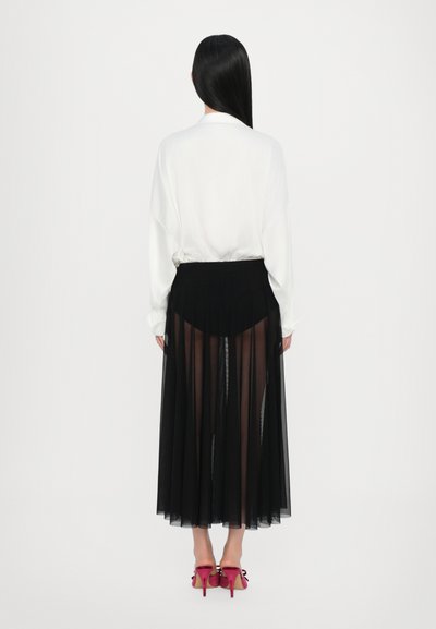 Camisa blanca de corte holgado, acompañada de una falda plisada negra y transparente. Zapatos de tacón negro con detalles en rosa completan el look.