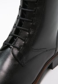 Botte en cuir noir à finition lisse, avec un design à lacets et une hauteur jusqu'à la cheville. Comprend des coutures verticales et un talon en bois.