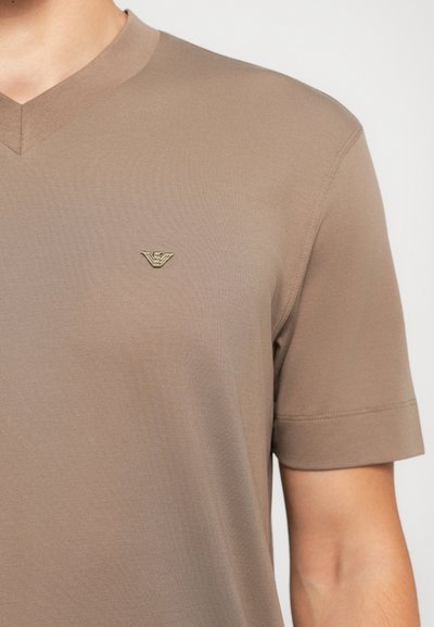Gros plan sur une personne portant un t-shirt beige à col en V et manches courtes, avec un petit logo d'aigle brodé sur la poitrine.