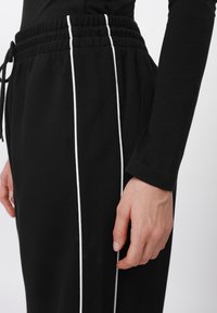 Fekete tréningnadrág fehér oldalsávokkal, elasztikus derékpánttal és húzózsinór részlettel, egy kéz lazán a szöveten nyugszik.