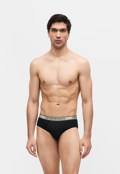 Νέος καλοντυμένος άνδρας σε όρθια στάση, που φοράει μαύρα εσώρουχα Emporio Armani με μπεζ ζώνη μέσης, μπροστά σε απλό ανοιχτό φόντο.