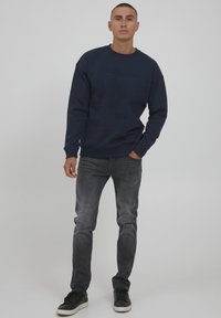 Felpa blu navy con collo rotondo, polsini e orlo a costine; abbinata a jeans grigi sbiaditi e sneakers nere con suole bianche.