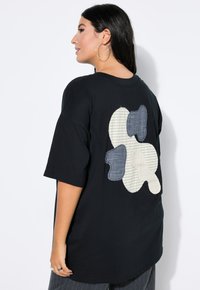 Sort oversized t-shirt med et tekstureret patchwork-design på bagsiden, med hvid, blå og stribet stofaccent. Korte ærmer.