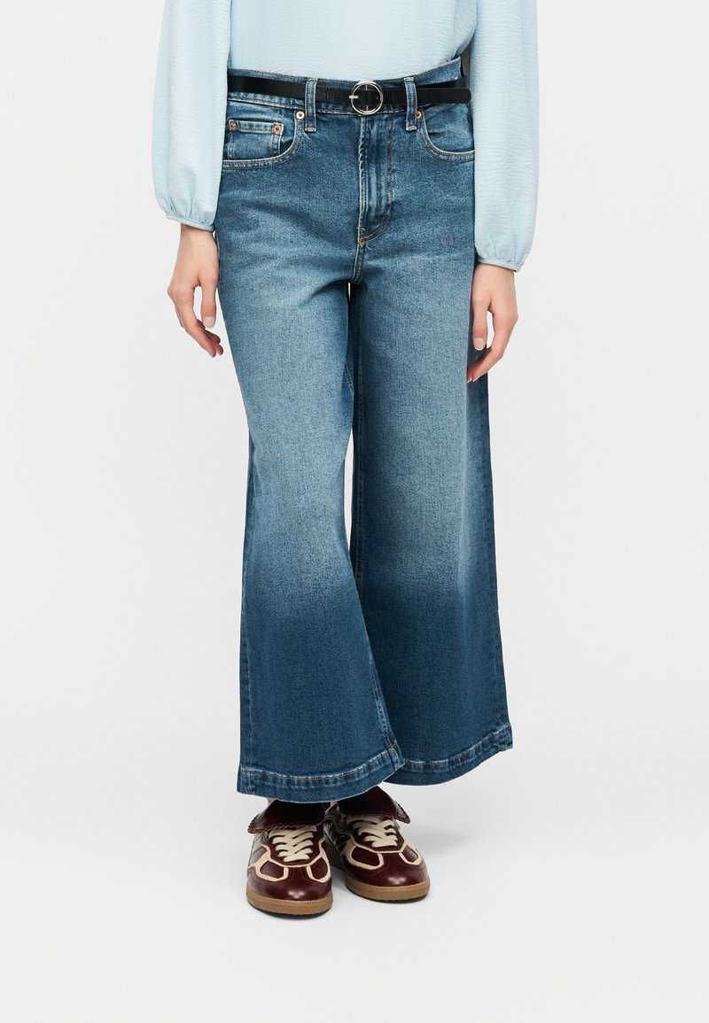 Άτομο που φοράει μπλε τζιν παντελόνι με ψηλή μέση και φαρδιά γραμμή, μαύρη ζώνη, ανοιχτό μπλε μπλούζα με μακριά μανίκια και μπορντό αθλητικά παπούτσια με λευκά κορδόνια.