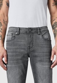 Hombre con vaqueros gris desgastado y una camiseta blanca de tirantes con canalé, mostrando tatuajes en ambos antebrazos y las manos relajadas a los lados.
