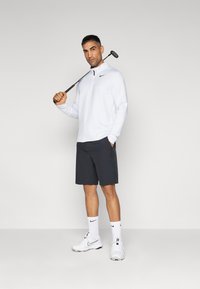 Pulover alb cu mânecă lungă și guler cu fermoar, pantaloni scurți negri, șosete sport albe și încălțăminte albă. Ținând un club de golf. Design simplu și athletic.