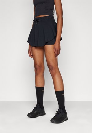Person iført sort atletisk nederdel med indbyggede shorts, sorte sokker og sorte løbesko, stående mod en ensartet lys baggrund.