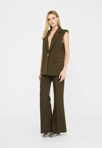 Gilet marrone su misura con ampi revers, chiusura a singolo bottone, abbinato a pantaloni a zampa d'elefante e scarpe con pattern. Tessuto dalla texture liscia.