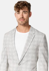Sacou gri deschis cu un model discret în carouri, guler cu umeri la piept, închidere cu un singur nasture și un buzunar pe piept, purtat peste un tricou alb.