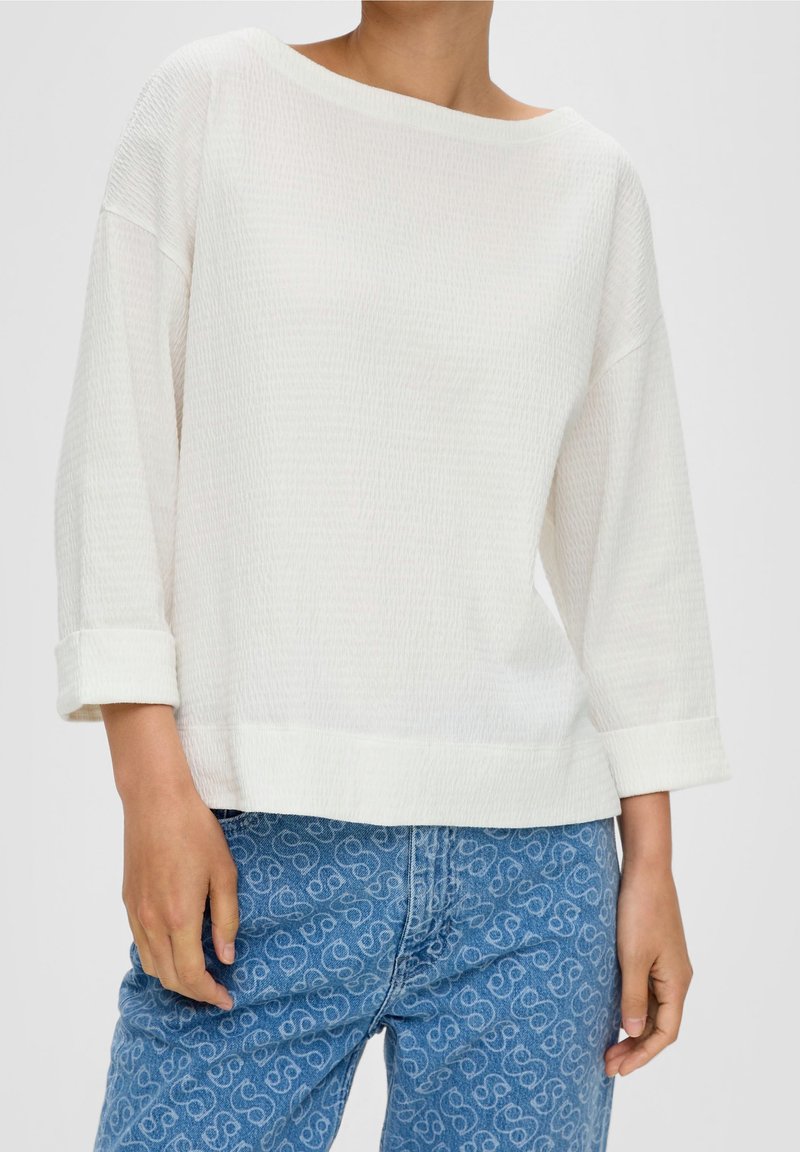 Valkoinen, teksturoitu, pitkähihainen paita, jossa on olkapäät paljastava kaula-aukko ja käärityt hihat, yhdistettynä sinisiin farkkuihin, joissa on hienovarainen kuviollinen suunnittelu.