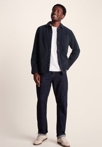 Dark blue corduroy shirt with button front, single chest pocket, layered over a white t-shirt, paired with dark jeans and beige sneakers.