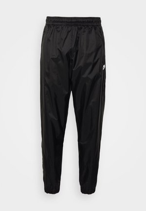 Black track pants made of lightweight, shiny material with an elastic waistband, tapered fit, and zippered pockets. Features a logo accent.
