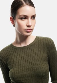 Maglione lavorato a maglia verde oliva con texture a coste e scollatura rotonda. La vista ravvicinata mette in risalto il motivo intricato e il materiale morbido.