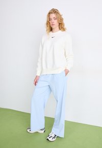 Sudadera crema con el logo negro de Nike, combinada con pantalones de pierna ancha en azul claro. El modelo lleva zapatillas blancas y negras. Fondo liso.