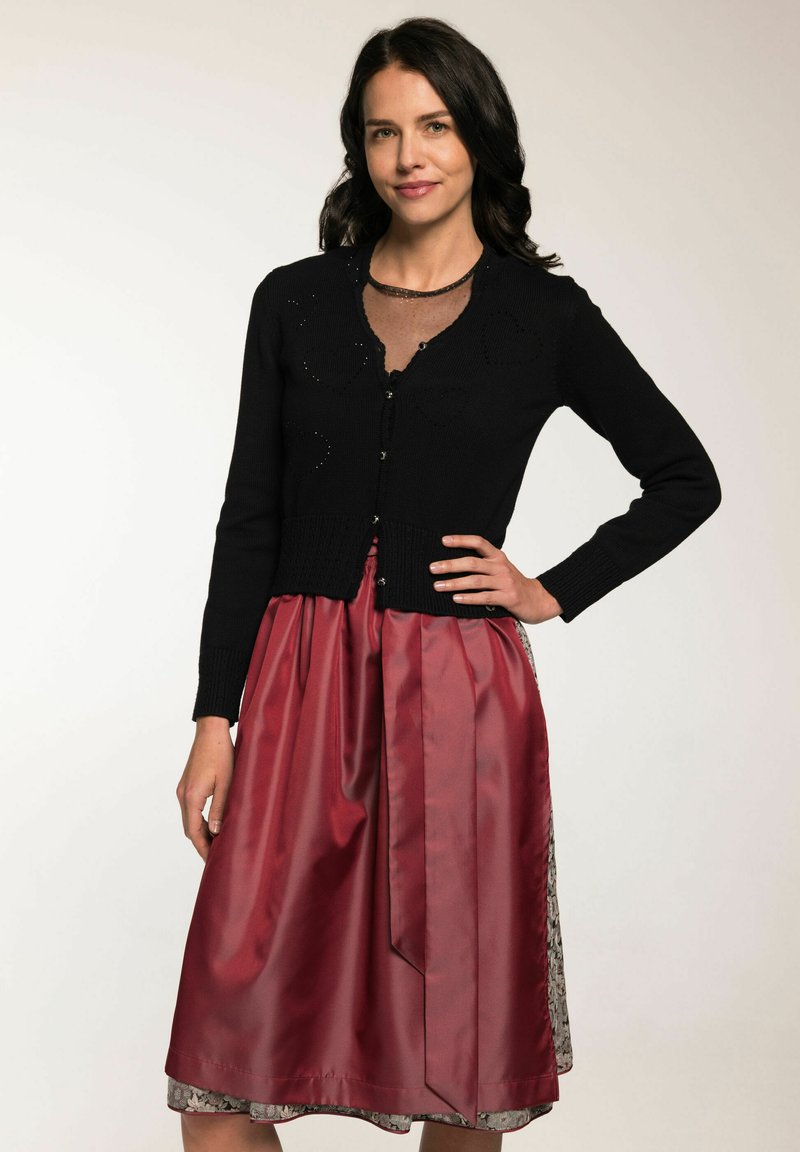 Schwarze Strickjacke mit V-Ausschnitt, ausgestattet mit einem strukturierten Muster und Knöpfen. Kombiniert mit einem glänzenden roten Satinrock, der mit einem gemusterten Saum akzentuiert ist.