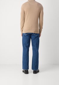 Uomo in piedi, che indossa un pullover beige a maniche lunghe, jeans dritti blu e scarpe nere, davanti a uno sfondo bianco semplice.