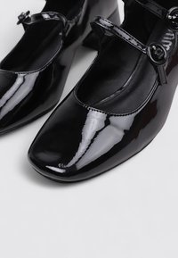 Chaussures Mary Jane en cuir verni noir avec bout rond et brides à bouton, présentées sur un fond clair uni.