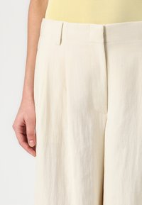 Cream-colored wide-leg trousers with pleats, featuring a smooth texture and side pockets. Waist has a clean finish with belt loops.