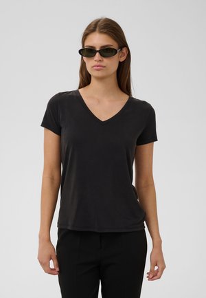 Donna con lunghi capelli castani che indossa una maglietta nera a maniche corte con scollo a V, pantaloni neri e occhiali da sole neri stretti, in piedi davanti a uno sfondo semplice.