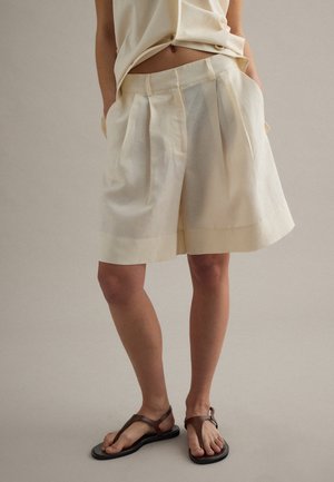 Persona vistiendo pantalones cortos crema sueltos de talle alto con pliegues y sandalias T de cuero marrón, de pie contra un fondo liso.