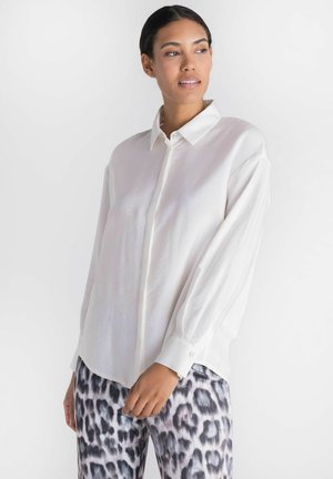 Femme portant une chemise blanche unie à manches longues boutonnée et un pantalon imprimé léopard noir et blanc, regardant sur le côté contre un fond blanc.