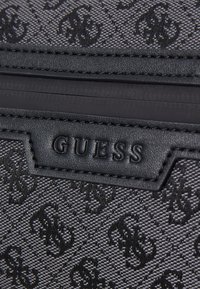 Black fabric wallet with embossed monogram pattern, featuring a textured surface, smooth leather accents, and a labeled black patch.