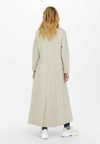 Abrigo largo beige con mangas largas, diseño recto, aberturas laterales, hecho de material suave, llevado sobre denim y zapatos blancos.