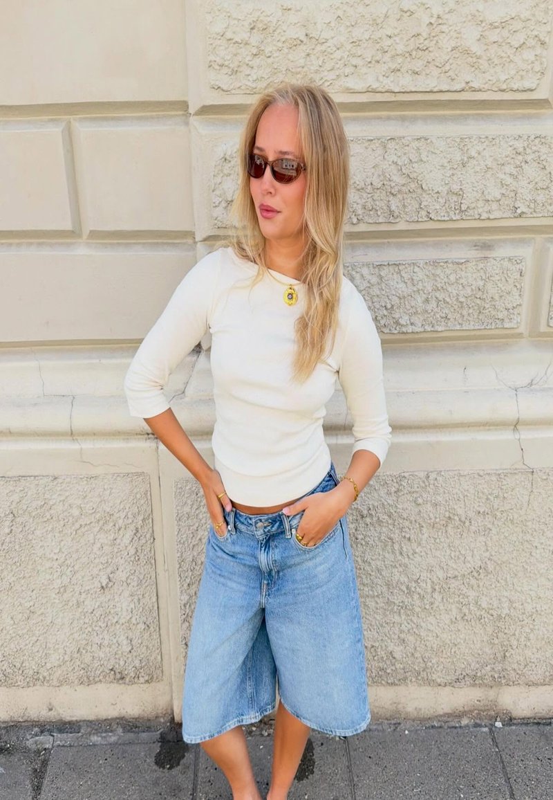 Γυναίκα με μακριά ξανθά μαλλιά, φοράει γυαλιά ηλίου, λευκό τοπ και φαρδιά μπλε τζιν σορτς, στέκεται μπροστά σε μια υφή λίθινο τοίχο.